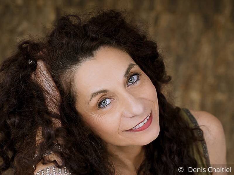 Portrait d'une femme brune aux cheveux ondulés, yeux bleus et qui sourit.